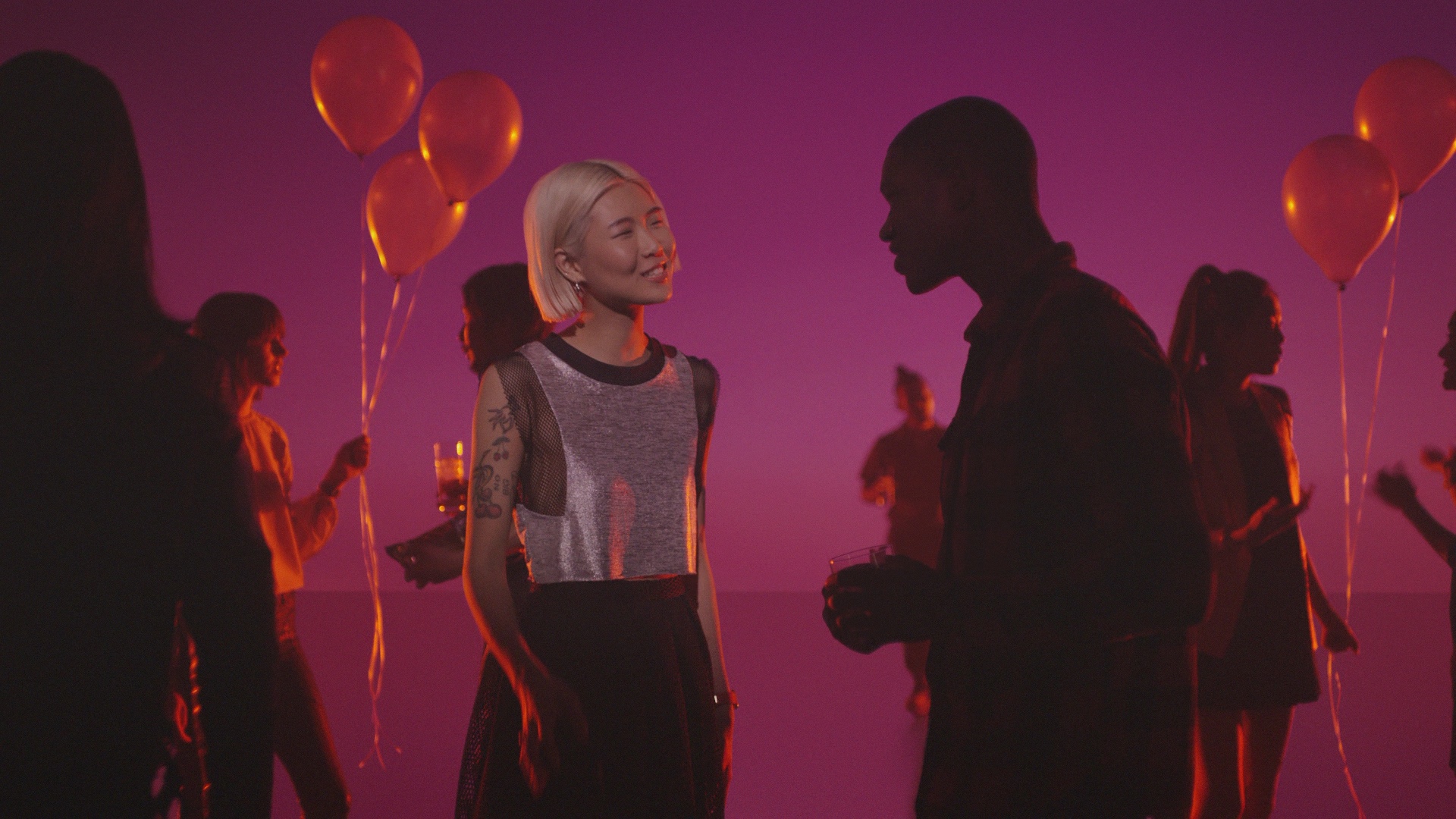 Party scene before adaptation, with a tattoo visible on the central performer’s upper arm.